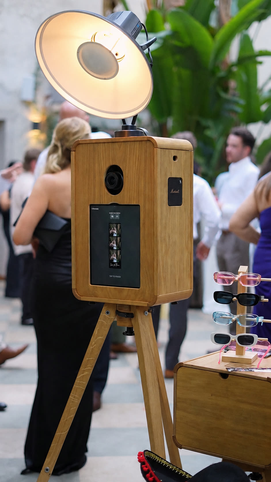 Fotomatón vintage de madera con flash de estudio en el patio de una boda, junto a la mesa de atrezzo con gafas y sombreros
