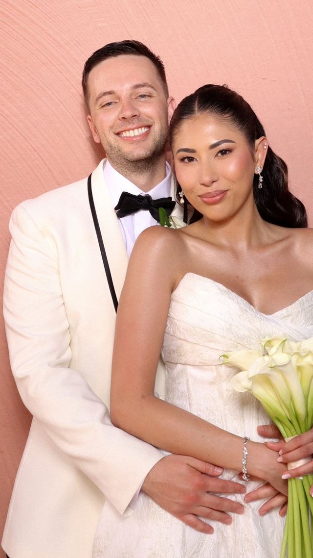 Novios sonrientes ante una pared rosa, él con esmoquin blanco y ella con un ramo de calas — fotomatón de boda
